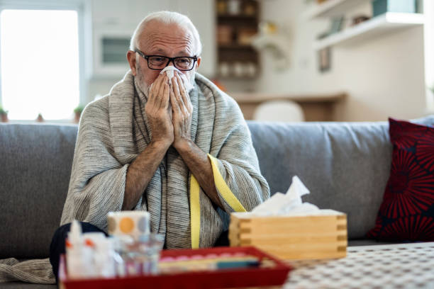 istock Senior man suffering from flu while sitting wrapped in a blanket on the sofa at home. Elderly Man blowing his nose while lying sick in bed at home. Man with a cold lying in sofa holding tissues. 1473505616