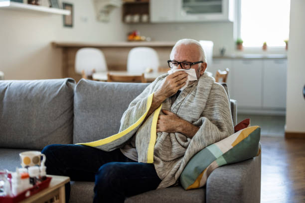 istock Cold exhausted senior man with flu wrapped in a warm blanket blowing his nose with a tissue in the livingroom. Man with a cold lying in sofa holding tissues. 1473505659