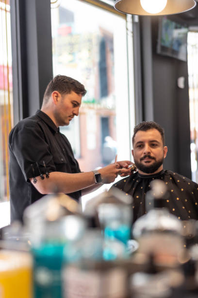 barbershop man gets a modern shave, beard and hairstyle, mirror reflection. parfum and products bottles blured - shaving men electric razor reflection imagens e fotografias de stock