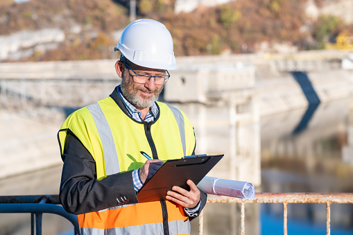 Bearded and bespectacled engineer in a blue suit, blue and white checkered shirt, orange and yellow safety reflective vest, and holding clipboard and pen in front of dam retaining wall.