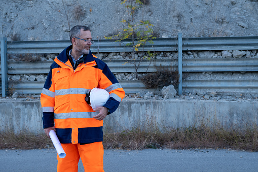 A bespectacled engineer in an orange-blue safety reflective suit with a blueprint in one hand and a white helmet under the arm of the other. He looks away.