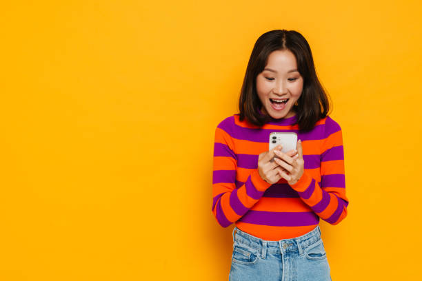 istock Excited asian girl using mobile phone isolated over yellow background 1473525562