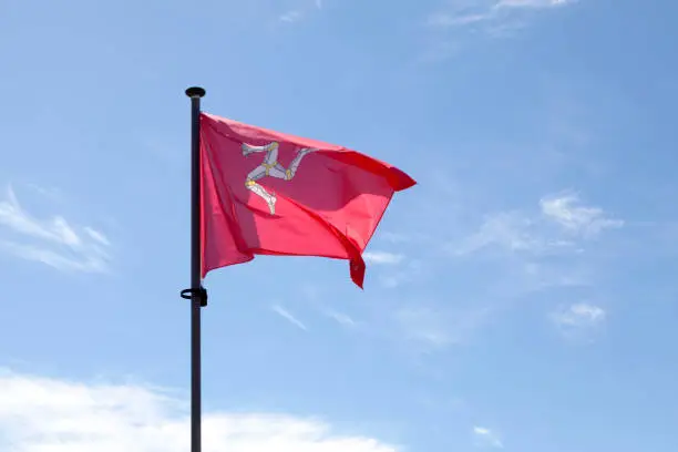Flag of Mann waving atop of its pole against a blue sky.