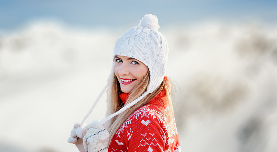 Young woman wear red winter pullover, white hat holding woollen braids with bobble ends, smiling, blurred snow covered mountains background