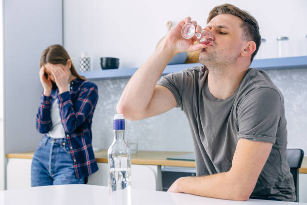 o homem bebe álcool por desespero, senta-se à mesa, está desempregado, preocupa-se com os problemas, ao fundo a esposa cobre o rosto para que a lágrima não seja visível. - abuso de álcool - fotografias e filmes do acervo