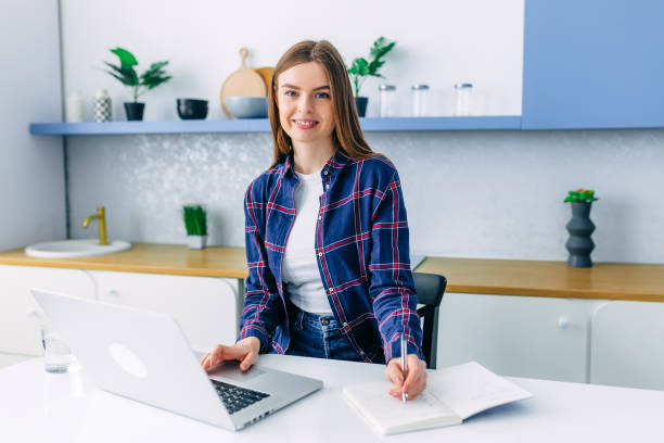 istock Young smiling happy beautiful woman relaxing using laptop computer, sits in the kitchen at the table at home. Young creative girl working laptop. Work from home. 1473528119