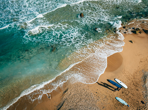 From an aerial perspective, surfing enthusiasts have finished their surfing activity and are walking forward with their surfboards in hand on a sandy beach where the sun is setting. They chat happily about their surfing experience and leave footprints on the sand.