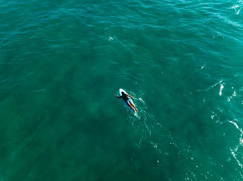 A male surfer is quietly lying on his surfboard in the transparent turquoise ocean, and from an aerial view, you can see him waiting for the waves to come, ready to take on the next surfing challenge at any moment.