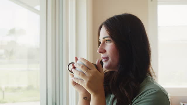 A young woman is drinking tea while she's looking through window