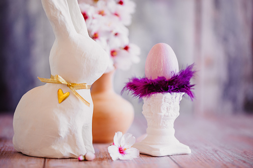 Beautiful close up of a purple painted Easter egg with lilac feathers on top of a small white pillar, a branch of almond blossoms and a white Easter bunny on blue vintage background. Creative color editing with added grain. Very soft and selective focus. Part of a series.