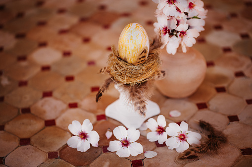 Gold painted Easter egg in a golden nest on top of a white pillar on an old outdoor stone table with almond blossoms. Creative color editing with added grain. Very soft and selective focus. Part of a series.