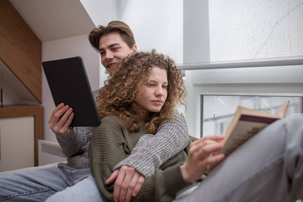istock Young woman reading book on cauch while her boyfriend is reading e-book 1473538934