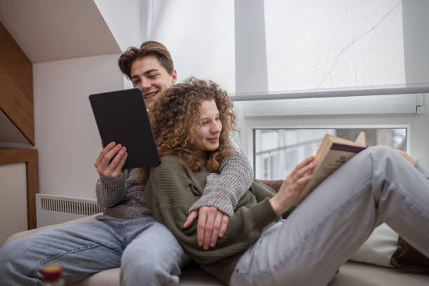 Young woman reading book on cauch while her boyfriend is reading e-book