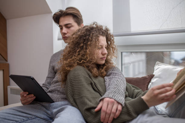 istock Young woman reading book on cauch while her boyfriend is reading e-book 1473538957