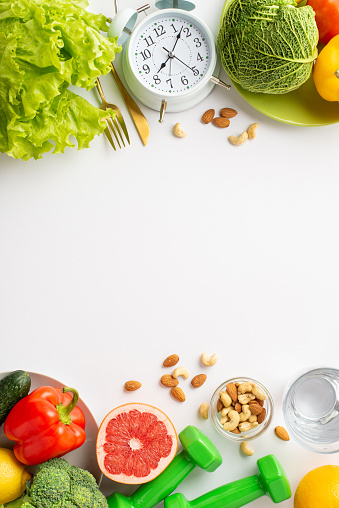 Proper diet concept. Top view vertical photo of plates with vegetables fruits nuts cutlery glass of water alarm clock and dumbbells on isolated white background with copyspace