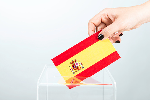 Human hand with black nail polish is inserting flag of Spain into ballot box. Representing elections in Spain.