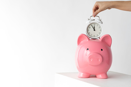 Hand of unrecognizable person is holding an alarm clock over pink piggy bank in front white background. The clock’s time is showing 5 to 12.