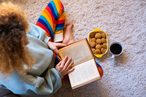 A woman with pajamas sitting on the floor at home when reading a book with hot drink and cookies.