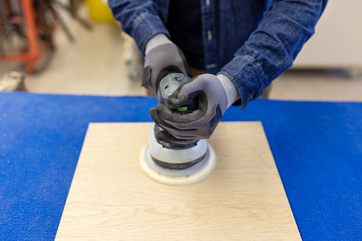 Close-up of a carpenter sanding a wood surface with orbital sander in workshop. Hands of an unrecognizable male worker using a sander on a piece of wood.