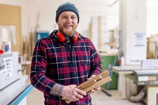 Portrait of carpenter with safety headphones and cap working at woodworking shop. Mid adult man holding wood pieces looking at camera and smiling in carpentry workshop.