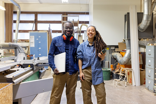 Portrait of two carpentry workshop workers smiling at camera. Male and female carpenter working in woodworking shop.