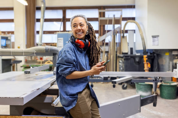 istock Woman holding a cell phone looking away and smiling at carpentry workshop 1473557868