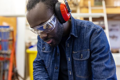 Close-up of a male carpenter wearing protective eyeglasses and headphones working in a carpentry workshop. African man working in a furniture factory with protective workwear.