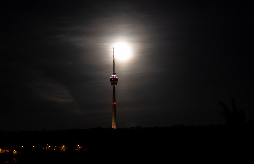 Full,  waning moon behind silhouette of the Stuttgart TV Tower. Hazy clouds around bright moon.