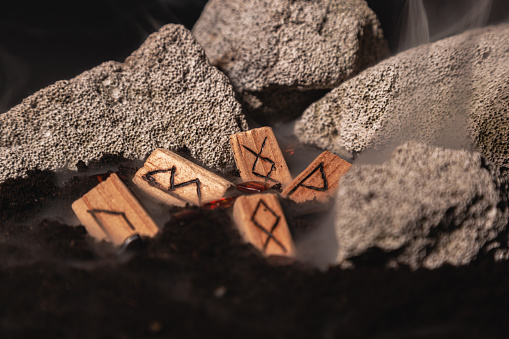 the wooden rune of Inguz among other runes, symbols of the earth, lie in the ground amid smoke and large stones. Mysticism and the rite of fruitful earth