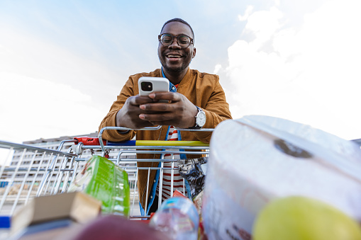 A smiling young African-American man holding a smart phone and coming back from groceries shopping
