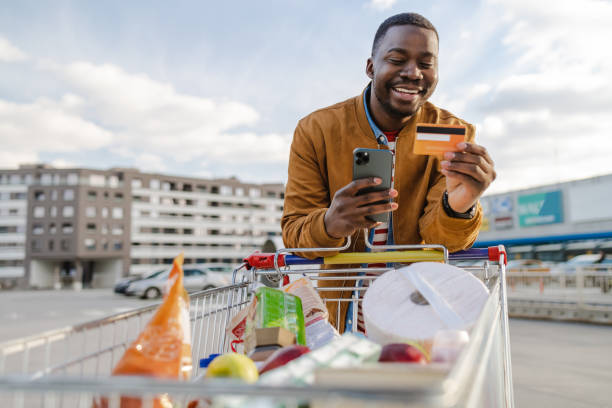 Portrait of a happy young African-American man pulling a shopping cart and using smart phone and credit card stock photo