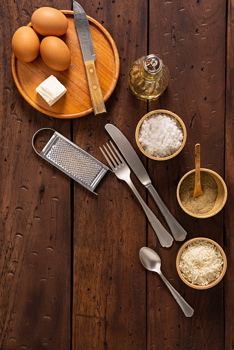 High angle view of rustic wood table in the kitchen at home. The food ingredients are eggs, cheese, back pepper, salt, cooking oil. Kitchen utensils are knife, spoon, scratcher. Ingredients is for a recipe to make avocado baked with eggs in the oven at home. But this image can be used for anything.