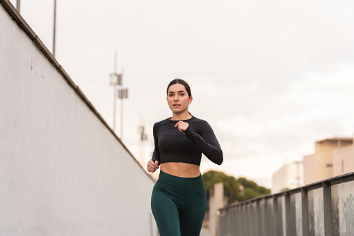 Medium shot of a young latina female athlete running towards the camera during her daily routine. She is wearing a dark grey long sleeve t-shirt and green leggings. With copy space