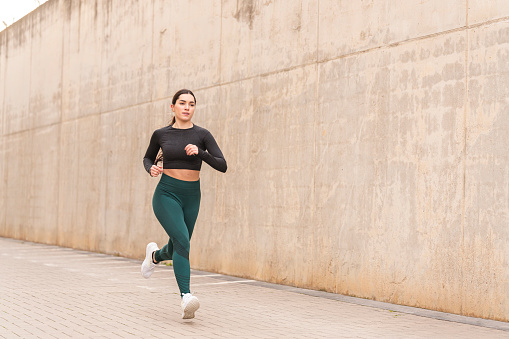 A focused young woman jogging outdoors in front of a concrete wall, following her daily physical activity routine. She has a fit and toned body and is wearing a dark grey long sleeve t-shirt and green leggings, full body image with copy space.