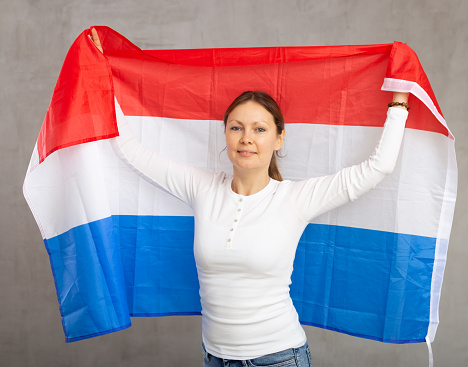 Smiling relaxed woman waving national flag of Netherlands, while looking at camera at gray wall background