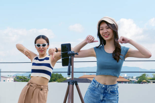 Young Asian woman with her friend tiktoker created her dancing video by smartphone camera together on rooftop outdoor at sunset. To share video to social media application stock photo