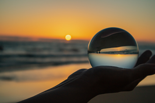 Unrecognizable female tourist demonstrating crystal sphere on blurred background of waving sea and sunset sky