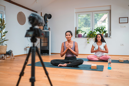 Attractive mixed race females starting a yoga class while filming with a professional camera. They are sitting on exercise mats with their hands together in prayer position.
