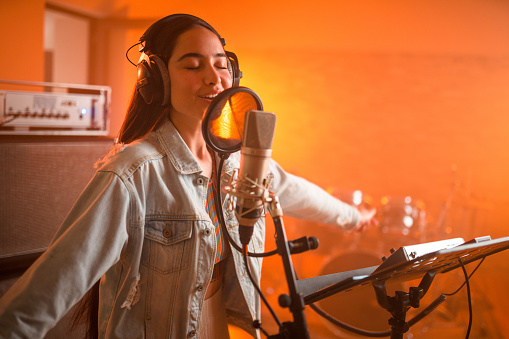 Young charming Asian female singer singing and recording songs in recording studio. Waist-up, eyes closed.