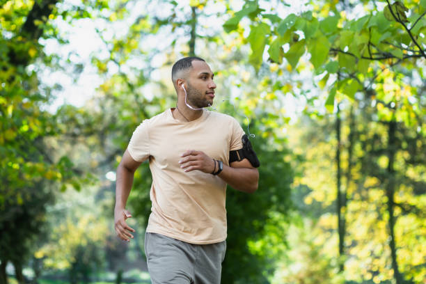 homme hispanique faisant du jogging dans le parc par une journée ensoleillée, coureur écoutant de la musique dans des écouteurs filaires, des livres audio et des podcasts, sportif heureux d’un week-end actif - black forest audio photos et images de collection