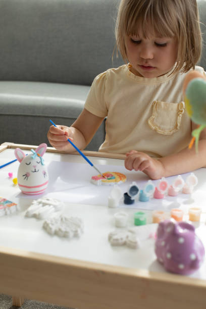 istock A cute little girl is sitting at a table and doing Easter crafts, coloring Easter figurines. Creativity and hobbies. 1473592815