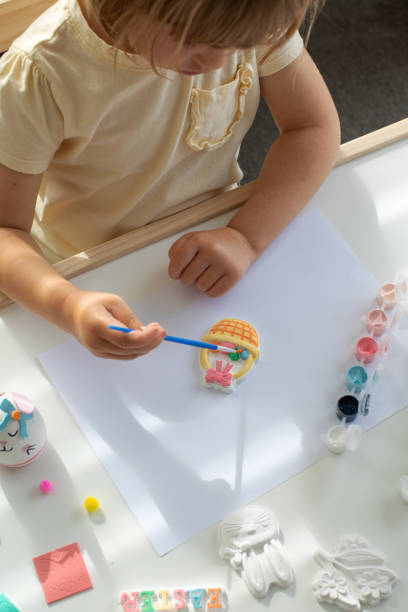 istock A cute little girl is sitting at a table and doing Easter crafts, coloring Easter figurines. Creativity and hobbies. 1473595466