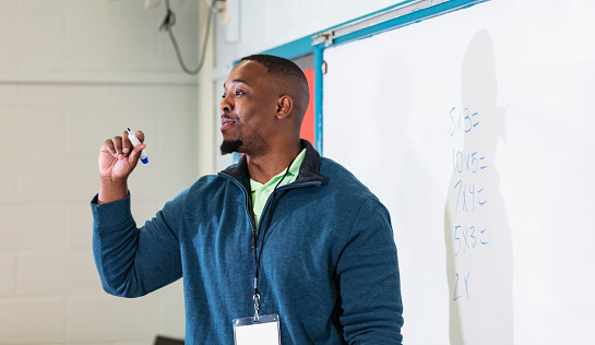 A young African-American teacher in the classroom teaching elementary school math. He is standing in front of a whiteboard holding a marker after writing equations. His back is to the board, facing his class.