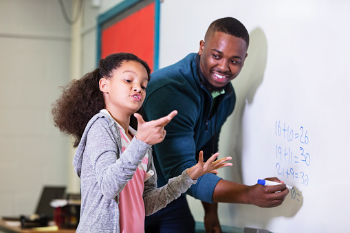 A multiracial 9 year old girl in math class, standing at the whiteboard with her teacher, a young African-American man in his 20s. The teacher is looking at his elementary student, waiting while she tries to figure out the answer. The focus is on the girl.
