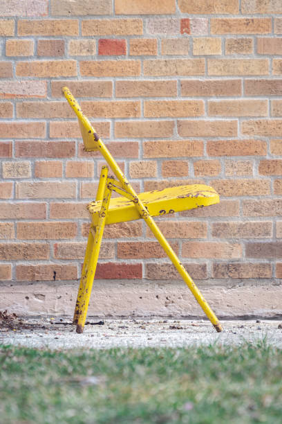 Side profile photograph of a bright yellow weathered and rusty metal folding chair sitting outside of a yellow brick building on pavement with grass in the foreground. stock photo