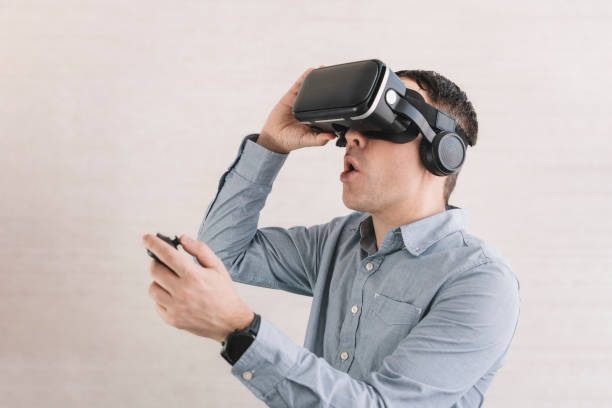 istock Young smiling man in VR glasses playing video game with joystick on a grey background, siting on the couch while having virtual reality experience, copy space 1473614452