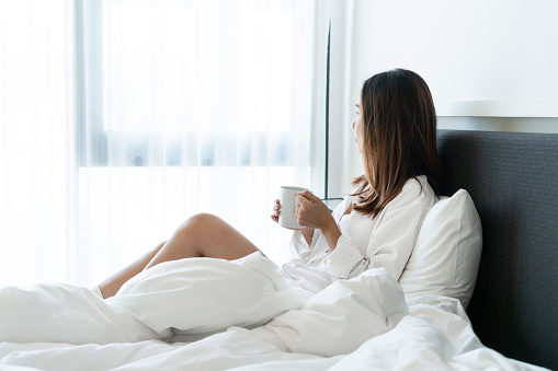 Young beautiful brunette hair woman in white shirt pajamas drinking coffee while sitting on the bed in the morning.