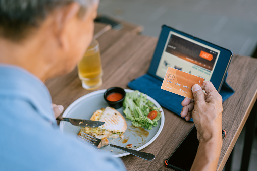 A senior Asian man is using his credit card and laptop to make online payments and shop at a cafe.