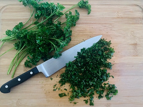 Horizontal flat lay of green fresh picked parsley from herb garden chopped up with knife on wood cutting board on domestic kitchen counter top