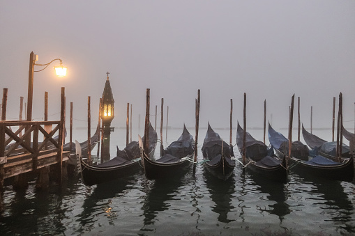 Secondary canal between residential buildings, small boats moored on the side, boat driving on water surface on a foggy day in Venice, Italy